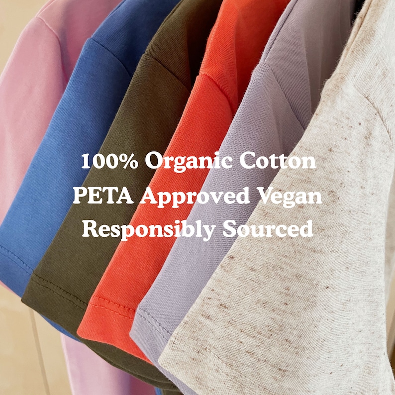 May include: A stack of six t-shirts in different colors, including pink, blue, green, red, purple, and beige. The shirts are made of 100% organic cotton and are PETA approved vegan. The text "100% Organic Cotton PETA Approved Vegan Responsibly Sourced" is printed on the shirts.