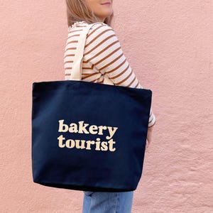 以下が含まれることがあります： 「bakery tourist」の文字がクリーム色のトートバッグ。バッグにはクリーム色のストラップが付いています。人物は茶色と白のストライプのトップスを着ています。背景はピンク色の壁です。