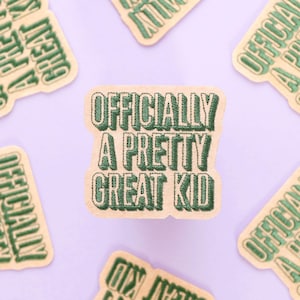 May include: Embroidered patch with the text "OFFICIALLY A PRETTY GREAT KID" in green on a tan background. Several similar patches are scattered around the central patch on a light purple surface.