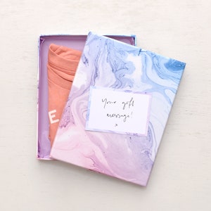 May include: A pink and blue marbled gift box with a handwritten note that says "Your gift message!" inside. The box is open and a folded pink shirt is visible inside.