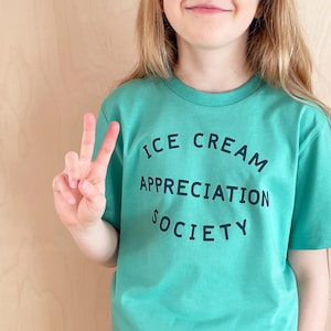 May include: A child wearing a teal blue t-shirt with the text "ICE CREAM APPRECIATION SOCIETY" printed in black.