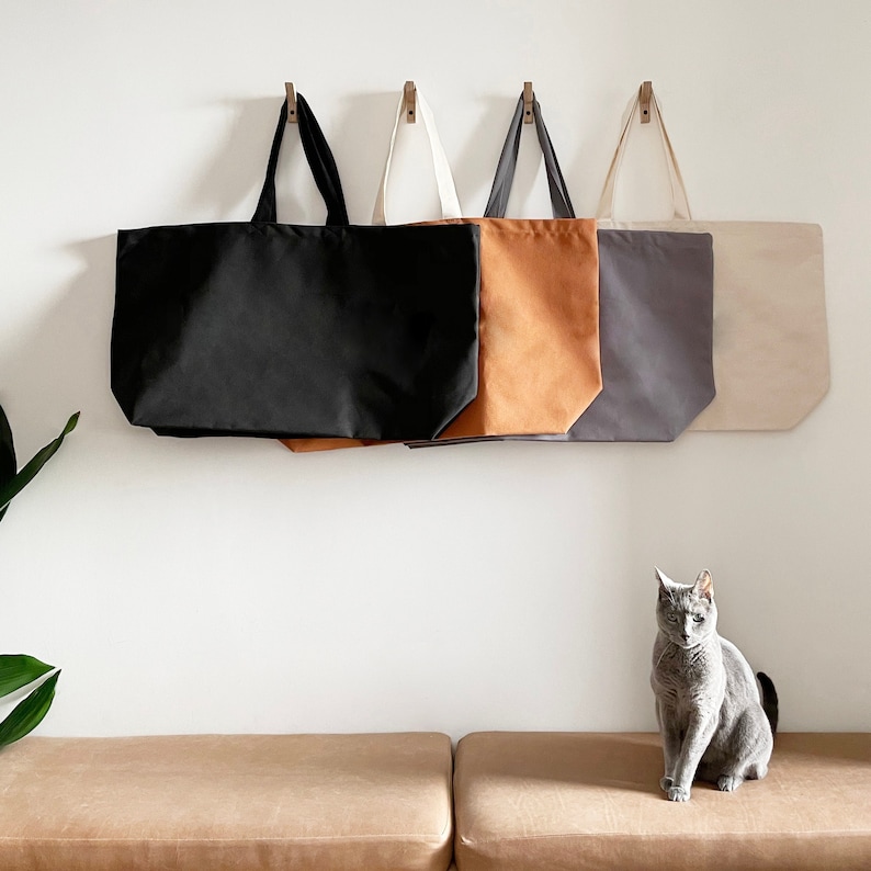 Op de afbeelding: Vier tote bags hangen aan haken tegen een witte muur. De tassen zijn zwart, bruin, grijs en wit. Een grijze kat zit op een bruine bank op de voorgrond.