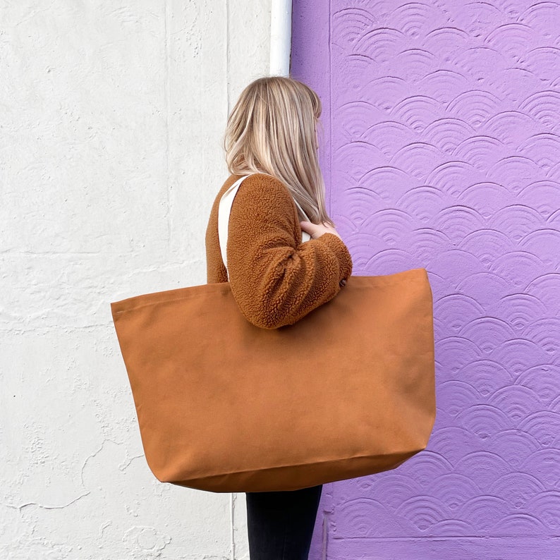 May include: A large brown canvas tote bag with white straps. The bag is being held by a person wearing a brown fuzzy jacket.