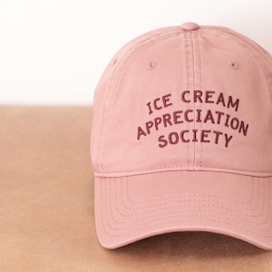 May include: A dusty rose baseball cap with the embroidered text "ICE CREAM APPRECIATION SOCIETY" in a darker rose color. The cap has a curved brim and a fabric strap closure.