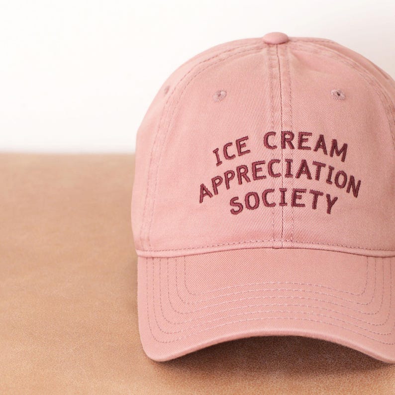 May include: A dusty rose baseball cap with the embroidered text "ICE CREAM APPRECIATION SOCIETY" in a darker rose color. The cap has a curved brim and a fabric strap closure.