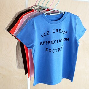 May include: A stack of t-shirts in various colors, including red, pink, blue, and gray. The top shirt is blue and has the text "ICE CREAM APPRECIATION SOCIETY" printed on it.