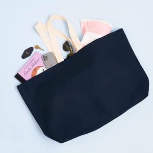 Op de afbeelding: Een donkerblauwe canvas tote bag met cr&egrave;mekleurige hengsels. De tas is gevuld met items waaronder een boek getiteld "Little House on the Prairie" van Laura Ingalls Wilder, een roze boek getiteld "Emmeline Pankhurst", een mobiele telefoon, een zonnebril en een roze-wit gestreepte handdoek.