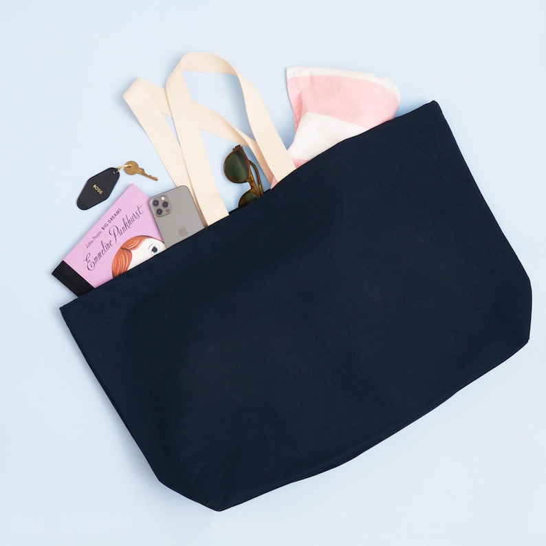 Op de afbeelding: Een donkerblauwe canvas tote bag met cr&egrave;mekleurige hengsels. De tas is gevuld met items waaronder een boek getiteld "Little House on the Prairie" van Laura Ingalls Wilder, een roze boek getiteld "Emmeline Pankhurst", een mobiele telefoon, een zonnebril en een roze-wit gestreepte handdoek.