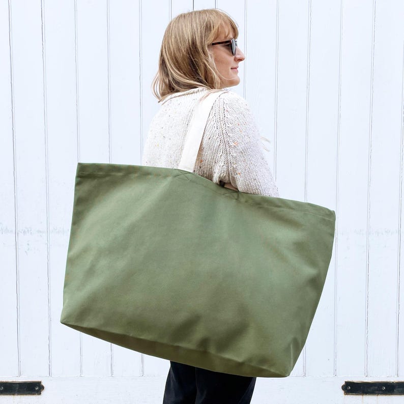 Op de afbeelding: Een grote, groene canvas tas met witte handvatten. De tas wordt gedragen door iemand die een witte trui draagt.