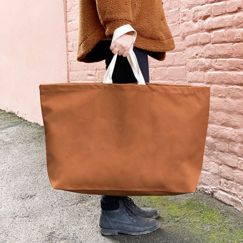 May include: A large brown canvas tote bag with white handles. The bag is being held by a person wearing a brown fuzzy jacket.