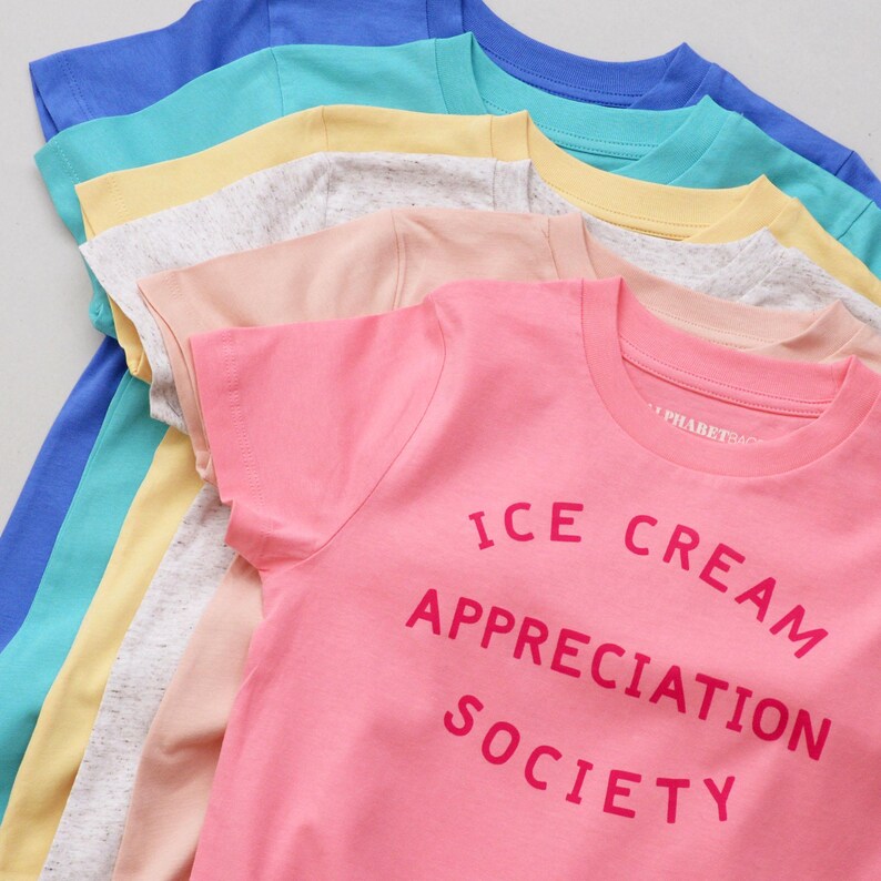 May include: A stack of colourful t-shirts with a pink t-shirt on top that says "ICE CREAM APPRECIATION SOCIETY" in pink lettering.