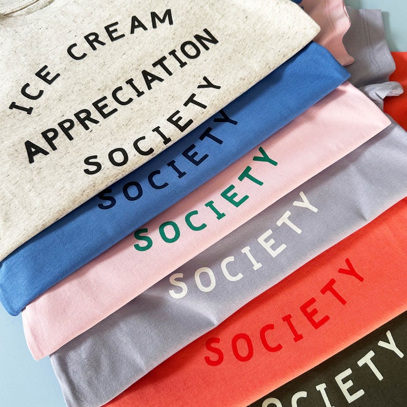 May include: A stack of t-shirts in various colors with the word "SOCIETY" printed on each shirt. The top shirt is beige with the words "ICE CREAM APPRECIATION SOCIETY" printed on it.