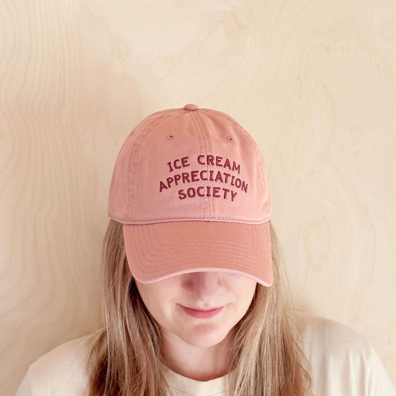 May include: A dusty rose baseball cap with the text "ICE CREAM APPRECIATION SOCIETY" embroidered in a darker rose color. The cap has a curved brim and a button on top. The hat is made of a soft, textured fabric.