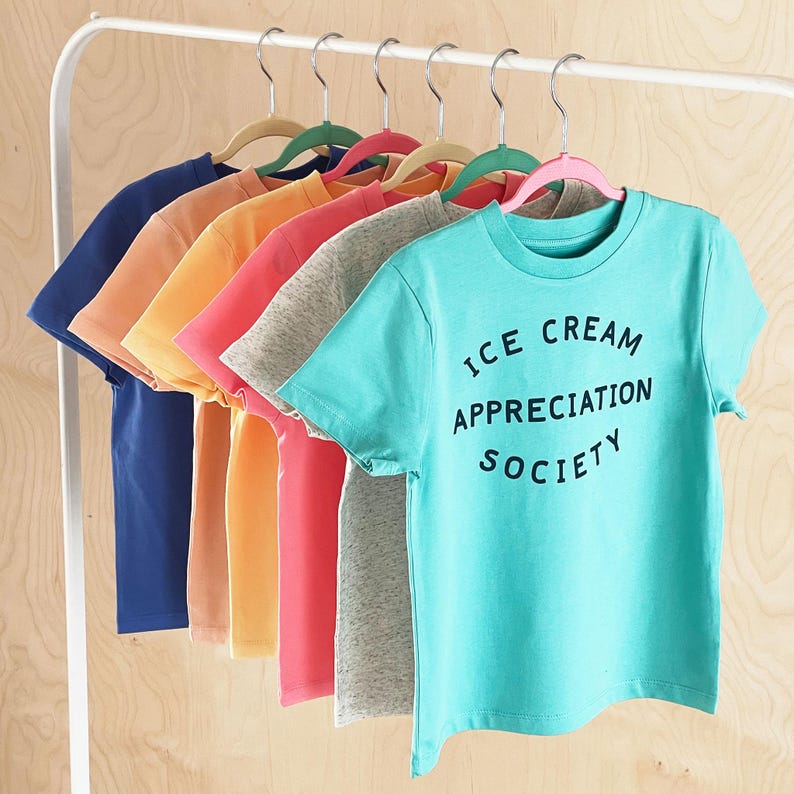 May include: A set of seven t-shirts hanging on hangers. The t-shirts are in various colours, including blue, orange, yellow, pink, grey, and turquoise. The turquoise t-shirt has the text "ICE CREAM APPRECIATION SOCIETY" printed on it.
