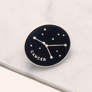 Puede incluir: Un pin de esmalte negro y blanco con la constelación del zodíaco Cáncer y la palabra "Cáncer" en letras blancas.