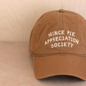 Könnte beinhalten: Braune Baseballkappe mit dem weißen Schriftzug "MINCE PIE APPRECIATION SOCIETY". Die Kappe hat einen gebogenen Schirm und einen Knopf oben. Der Hut ist aus weichem, abgenutztem Material.