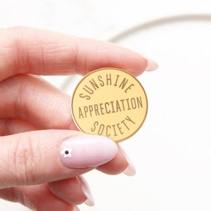 Pode incluir: Um alfinete redondo dourado com o texto "Sunshine Appreciation Society" em letras marrons.