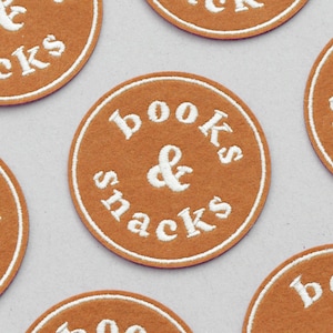 Puede incluir: Un parche redondo de fieltro marrón con bordado blanco que dice "books & snacks".