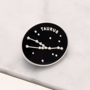 May include: A black enamel pin with a silver border featuring the Taurus constellation and the word "TAURUS" in white lettering.