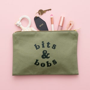 May include: A green canvas zippered pouch with the text "bits & bobs" printed in black. The pouch is open and contains a variety of items, including a key chain, a lipstick, a makeup brush, and a small toy.