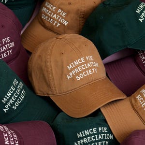 Könnte beinhalten: Eine Sammlung von Baseballkappen in verschiedenen Farben, darunter Braun, Grün und Burgunder. Jede Kappe trägt den weißen Text "MINCE PIE APPRECIATION SOCIETY" auf der Vorderseite. Die Kappen sind aus robustem Stoff gefertigt und haben einen klassischen, gebogenen Schirm.