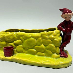 May include: A ceramic planter featuring a yellow stone wall design with a red bucket on the left side and a red elf figure leaning against the right side. The base is yellow and textured to resemble a stone path, with a hollow center for planting.
