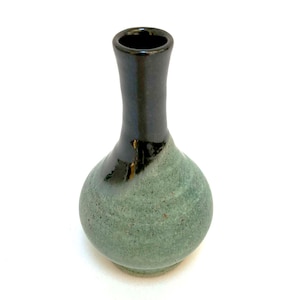May include: A small, handcrafted ceramic vase with a rounded body and a slender neck. The vase features a two-tone design, with a dark, glossy black top and a textured, speckled green base. The vase is suitable for displaying small flowers.