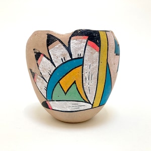 May include: A small, hand-painted ceramic pot with a heart shape. The pot is decorated with a colorful geometric design featuring white, black, blue, green, yellow, and pink.