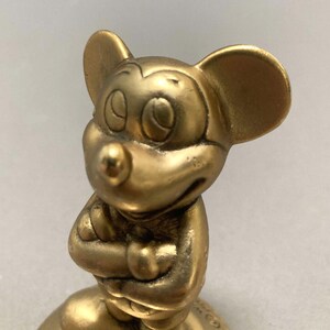 Op de afbeelding: Een gouden Mickey Mouse-figuur met gekruiste armen, staand op een ronde basis. Het figuur is gemaakt van een glad, glanzend materiaal.