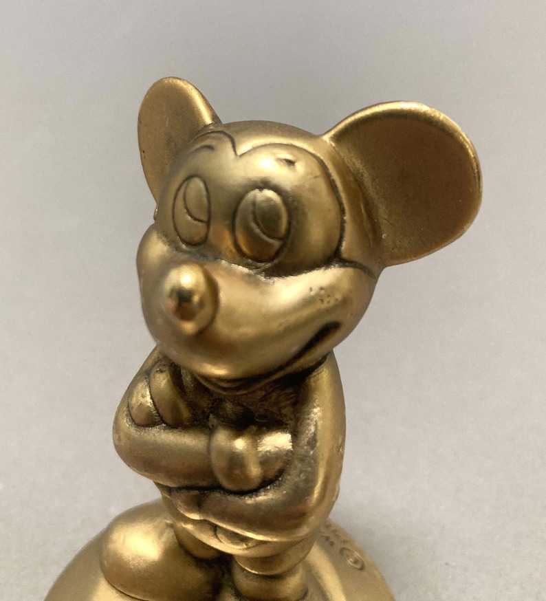 Op de afbeelding: Een gouden Mickey Mouse-figuur met gekruiste armen, staand op een ronde basis. Het figuur is gemaakt van een glad, glanzend materiaal.