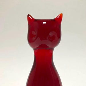 May include: A red glass cat-shaped vase with a glossy finish. The vase features a slender neck that widens to a rounded body and a head with pointed ears and circular eye indentations. The overall design is minimalist and modern.