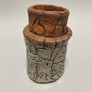 May include: A small, hand-crafted ceramic vase with a textured, brown top and a white bottom with a carved design.