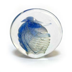 May include: A clear glass paperweight with a blue and white bird design encased inside. The bird has detailed wings and a beak, with air bubbles visible within the glass. The paperweight is round and sits on a white surface.