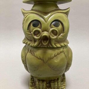 May include: A green ceramic owl figurine wearing a graduation cap and glasses. The owl has large, round eyes and a wide, open beak. The figurine is standing on a square base.