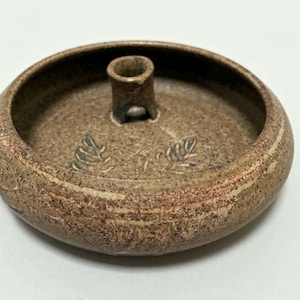 May include: A handcrafted, brown ceramic candle holder with a circular base and a central, cylindrical support. The holder features leaf-like etchings and a textured surface, perfect for home decor.