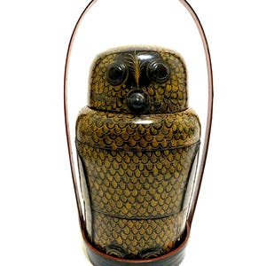 May include: A black and gold owl-shaped bento box with a handle. The box is decorated with a scale pattern and has a dark brown leather handle. The owl's eyes and beak are prominent features. The bento box is a unique and decorative item.