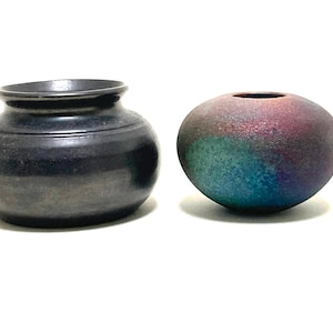 May include: Two ceramic vases. One is a dark, matte black, squat pot with a wide opening. The other is a round, spherical vase with a color gradient of purple, teal, and blue. Both vases are on a white background.