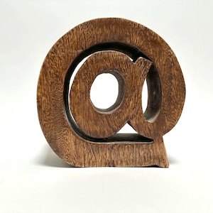 May include: A freestanding wooden sculpture shaped like the at symbol. The object features a natural brown wood grain pattern with a smooth finish, carved into a circular form with a central loop and a tail that curves around the exterior.