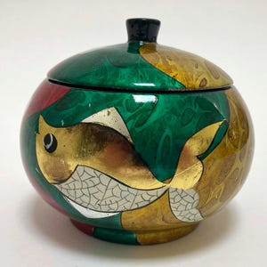May include: A round, lidded decorative box with a fish design. The box is painted with a green, gold, red, and black color scheme. The fish is gold with white and silver details. The lid has a small black knob.