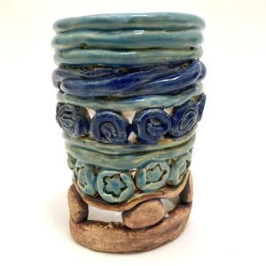 May include: A handmade ceramic vase with a cylindrical shape. The vase features bands of varying textures and colors, including shades of blue and brown. The design incorporates spiral and floral patterns, creating a unique, artistic appearance.