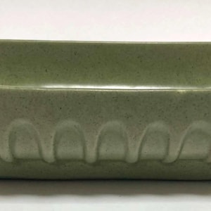 May include: A rectangular, sage green ceramic dish with a speckled texture. The dish has a slightly flared rim and a row of arched, decorative details along the lower edge. The corners are gently angled.