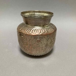 May include: A silver-colored metal pot with a wide body and a narrow neck. The pot features intricate embossed designs, including figures and floral patterns. A band of copper-colored metal encircles the middle of the pot.