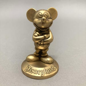 Op de afbeelding: Een gouden Mickey Mouse-figuur met gekruiste armen, staand op een ronde basis met het woord "Disneyland" erop.
