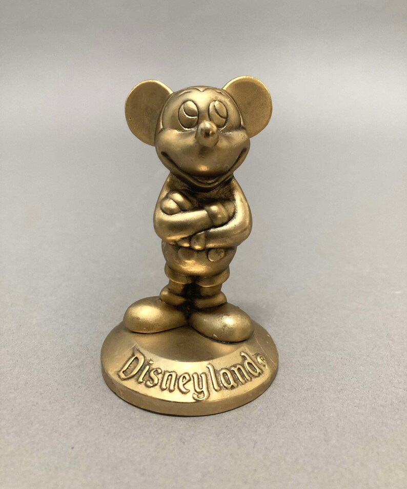 Op de afbeelding: Een gouden Mickey Mouse-figuur met gekruiste armen, staand op een ronde basis met het woord "Disneyland" erop.