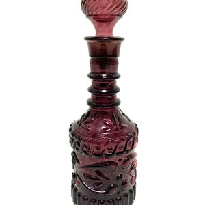 May include: A vintage, amethyst-colored glass decanter with a decorative stopper. The bottle features an ornate, embossed design and a ribbed stopper. The decanter is approximately 10 inches tall.