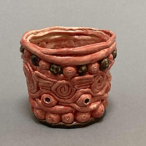 May include: A small, pink ceramic pot with a textured, patterned design. The pot has a brown rim and is decorated with small, brown beads.