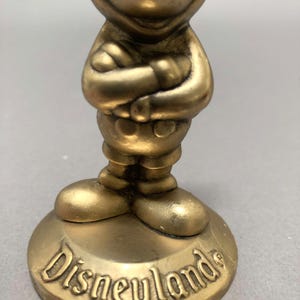 Op de afbeelding: Een gouden Mickey Mouse-figuur met gekruiste armen, staand op een ronde basis met het woord "Disneyland" erop.