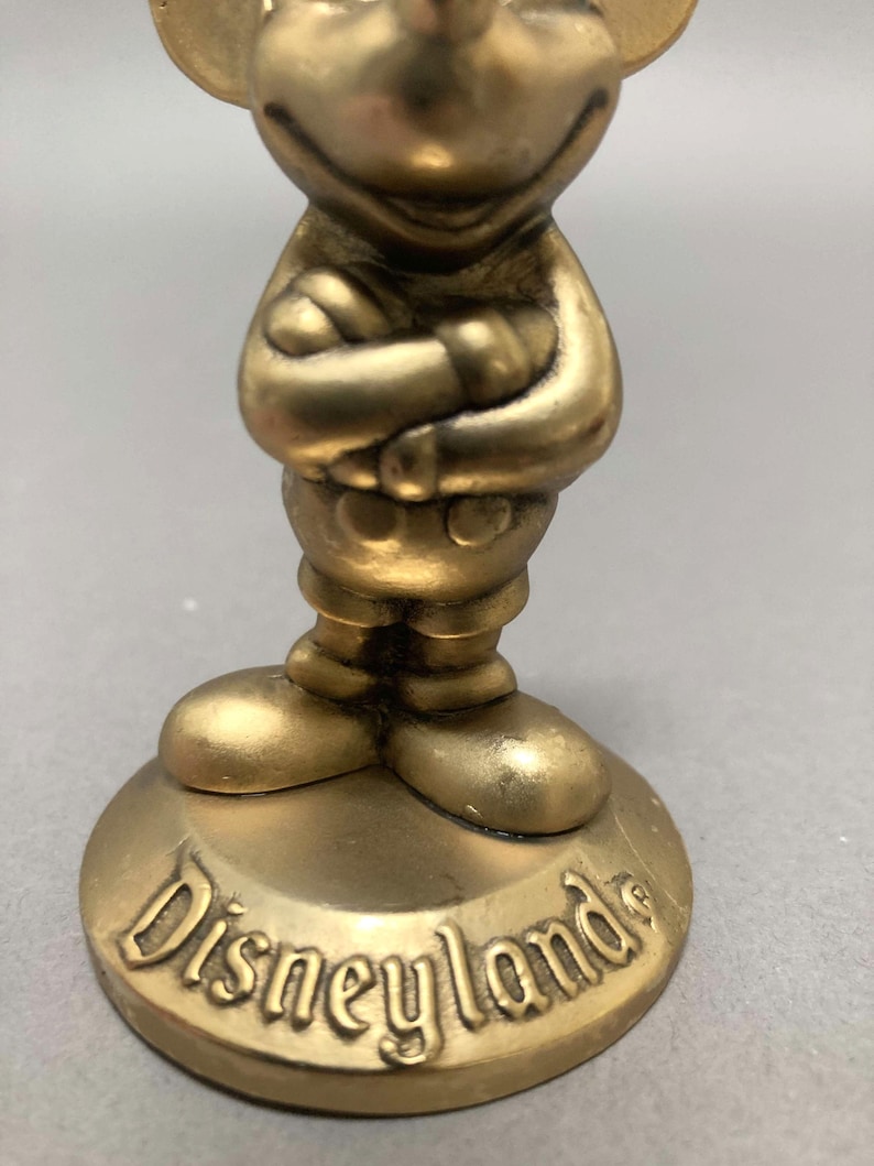 Op de afbeelding: Een gouden Mickey Mouse-figuur met gekruiste armen, staand op een ronde basis met het woord "Disneyland" erop.