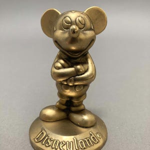 Op de afbeelding: Een gouden Mickey Mouse-figuur met gekruiste armen, staand op een ronde basis met het woord "Disneyland" erop.
