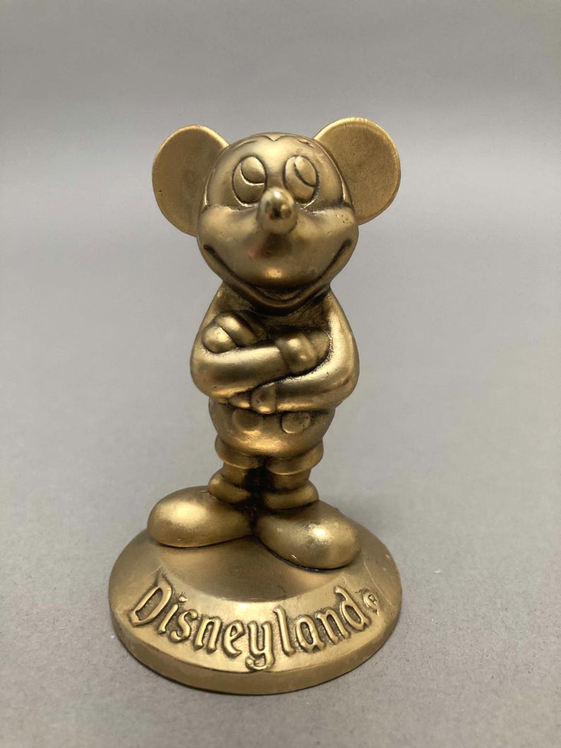 Op de afbeelding: Een gouden Mickey Mouse-figuur met gekruiste armen, staand op een ronde basis met het woord "Disneyland" erop.